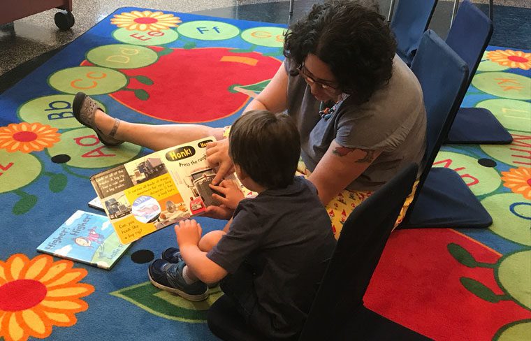 An adult interacts with a child over a book on a colorful rug