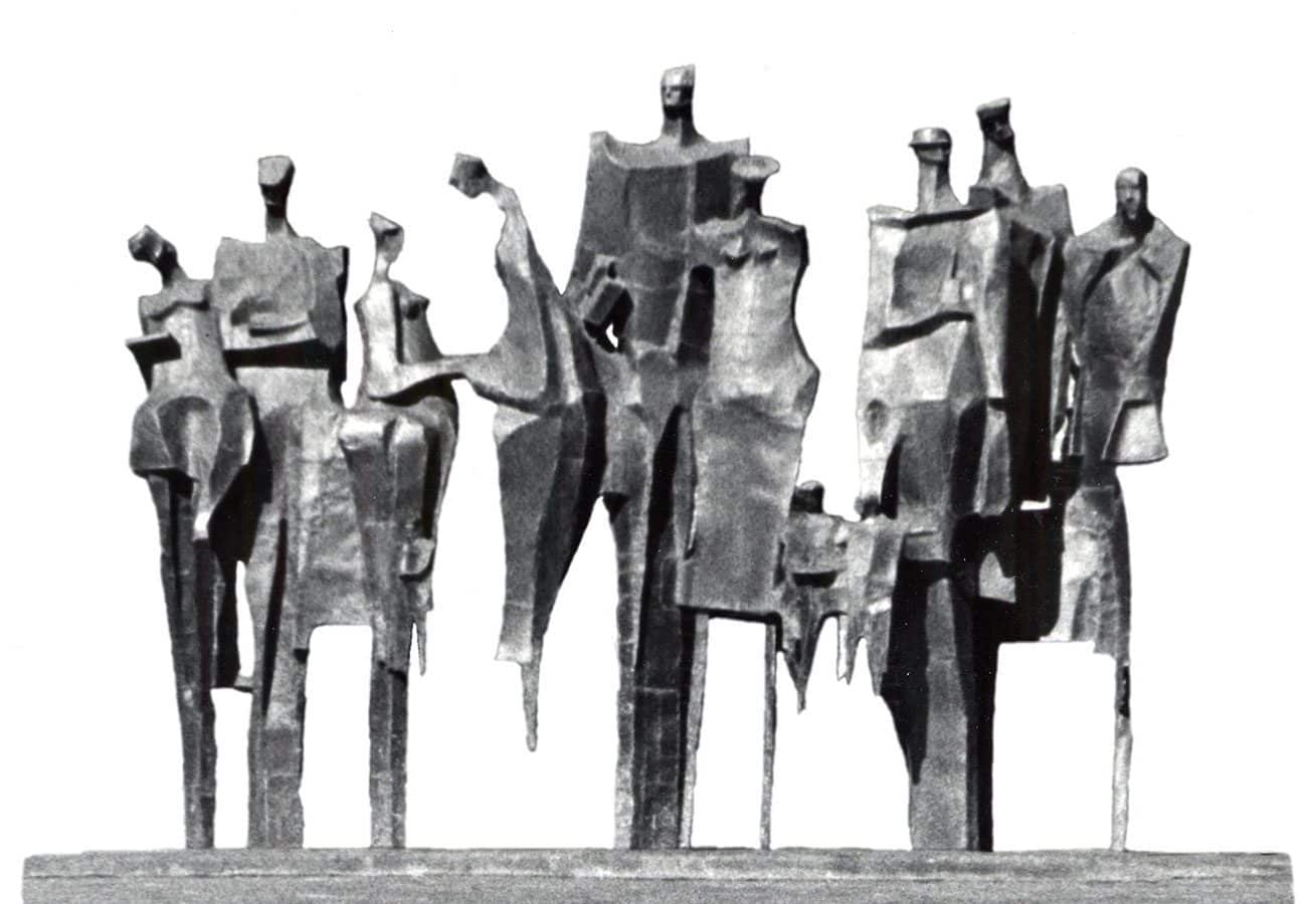 Twelve angular human figures stand in a row in this large-scale statue. Some figures face outward in various directions while others appear to engage with one another.