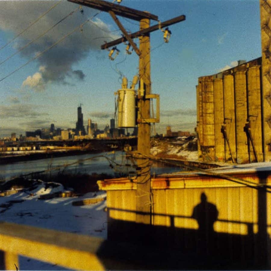A view of the Chicago skyline in the background with the shadow of a person standing on a bridge projected onto an industrial building and an electrical pole in the foreground, and water show in the middle ground.