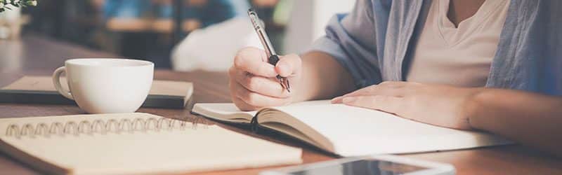 Person writing on a notebook beside teacup and tablet computer