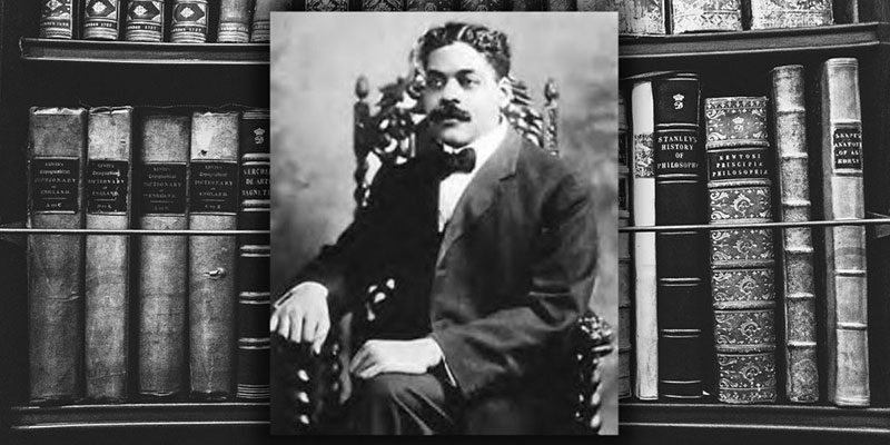 A portrait of Arturo Alfonso Schomburg sitting in a chair with books on a shelf in the background