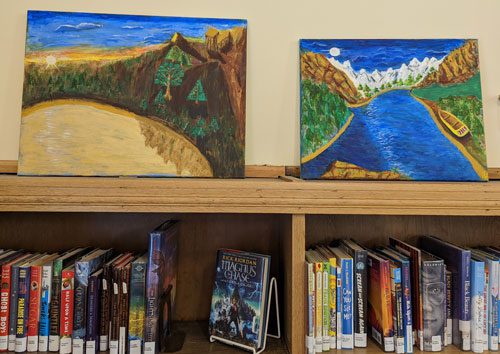 Two paintings above a bookshelf. One shows a sunset over a lake, surrounded by trees and mountains. The other shows a river valley between snowy mountains and a wooden boat on the shore.
