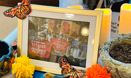 Framed picture of protest against attacks on Latin American environmental activists, set on a Dia de Muertos ofrenda with candles, paper marigolds, and paper monarch butterflies