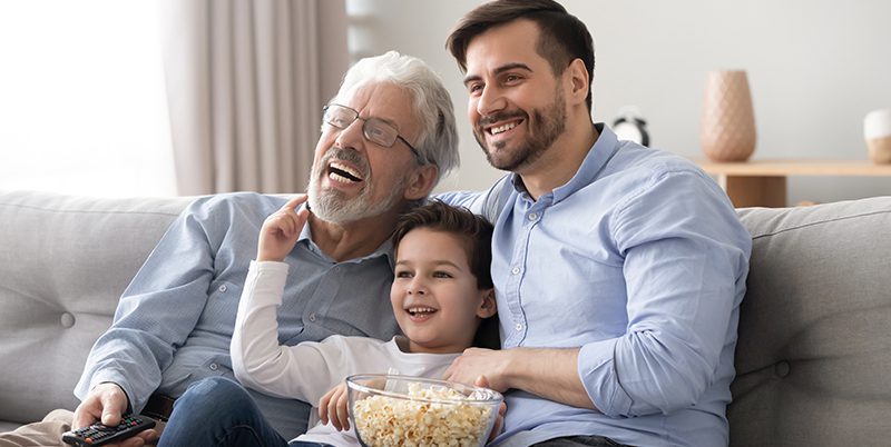 A grandfather, dad, and son laughing and eating popcorn while watching a movie together on the couch