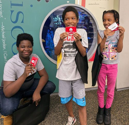 Three kids pose with their library cards in front of the Main Library aquarium