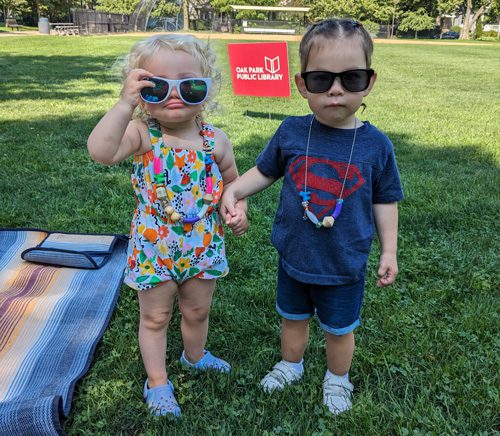 Two toddlers hold hands, wear sunglasses and summer reading bead necklaces outside at a park