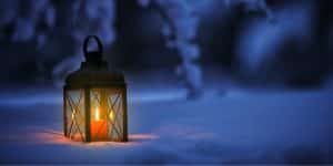 A lantern with a glowing candle inside sitting in the snow under a tree