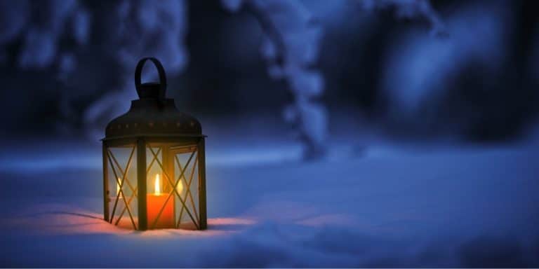 A lantern with a glowing candle inside sitting in the snow under a tree