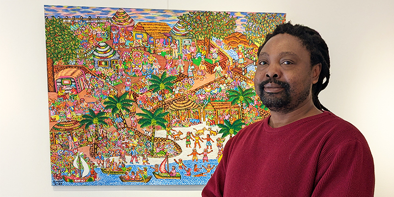 The artist poses next to his painting, an intricate, colorful scene of a village.