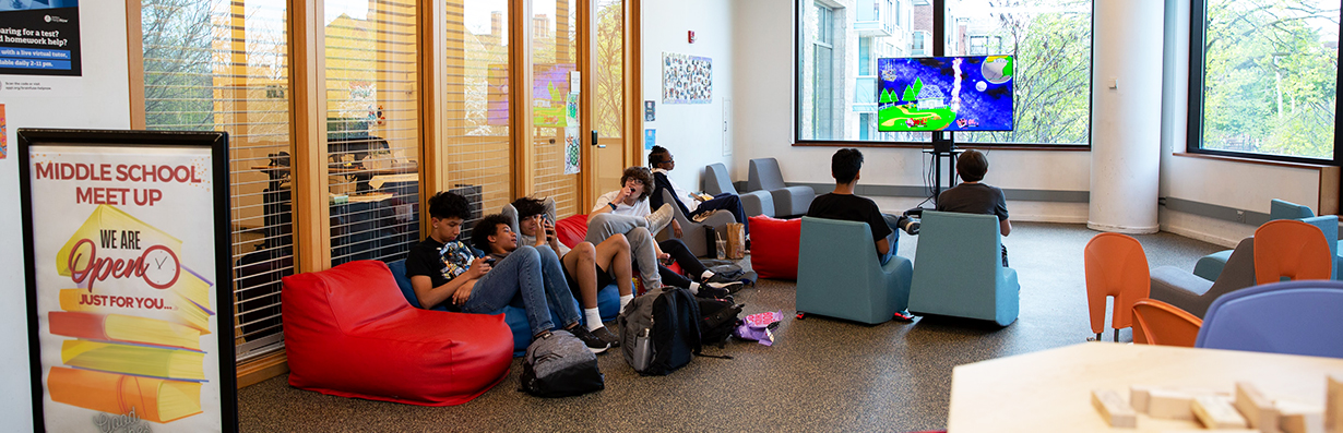 Middle schoolers sitting on bean bags and gaming chairs, playing video games, chatting, and hanging out in the Main Library
