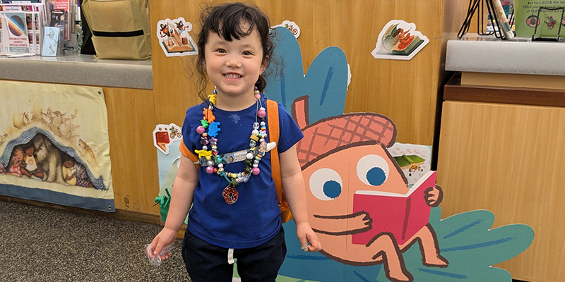 Child smiles and poses next to a library desk. She's wearing lots of summer reading beads on necklaces and behind her is a large cardboard cutout of an acorn reading a book on an oak leaf.