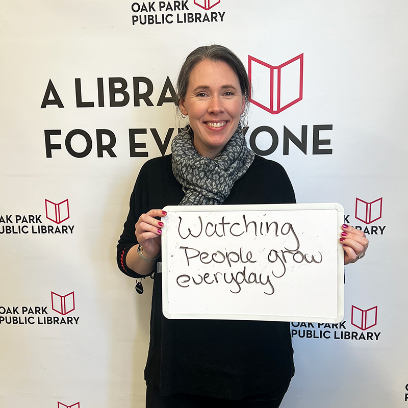 Library staff member holding a sign that reads, Watching people grow every day