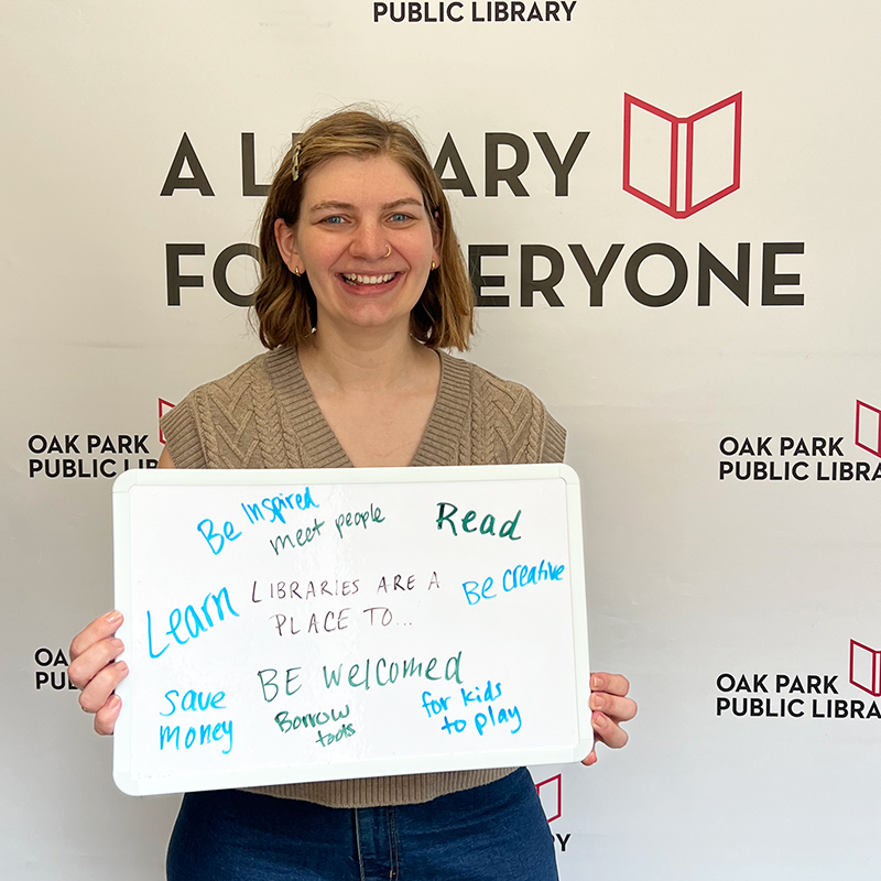 Library staff member holding a sign that reads, Libraries are a place to be: inspired, meet people, read, be creative, learn, save money, be welcomed, borrow tools, for kids to play.