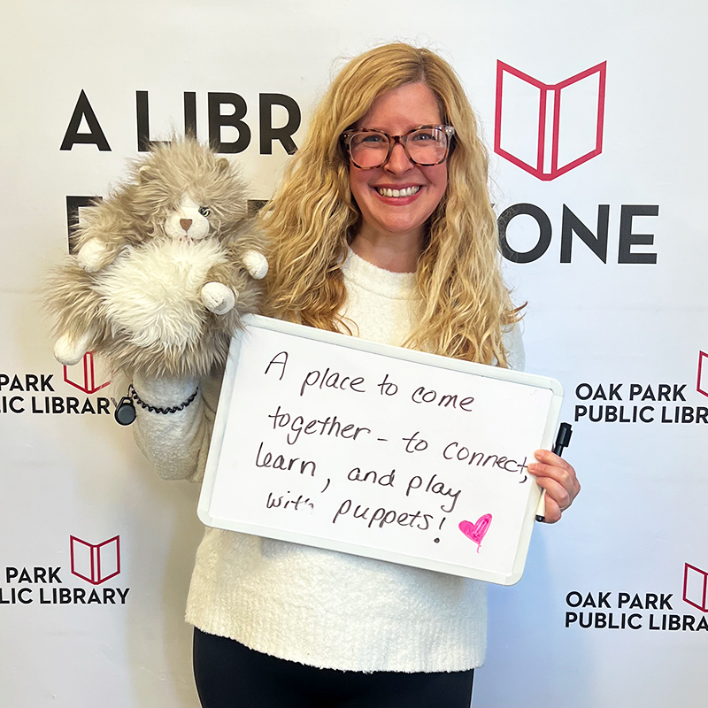 Library staff member holding a sign that reads, A place to come together—to connect, learn, and play with puppets!