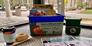 In a cafe window, there's a tray with a to-go coffee cup and a sesame seed bagel, a bin of kids books with a sign that says "Read while you wait," and paper event calendars