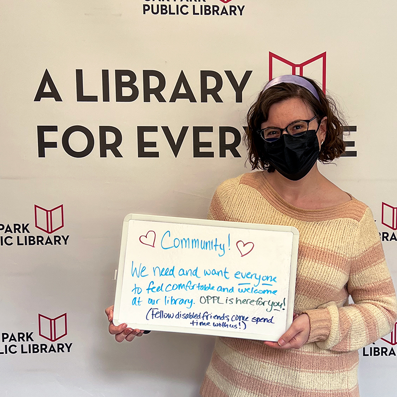 Library staff member holding a sign that reads, Community! We need and want everyone to feel comfortable and welcome at our library. OPPL is here for you! (Fellow disabled friends, come spend time with us!)