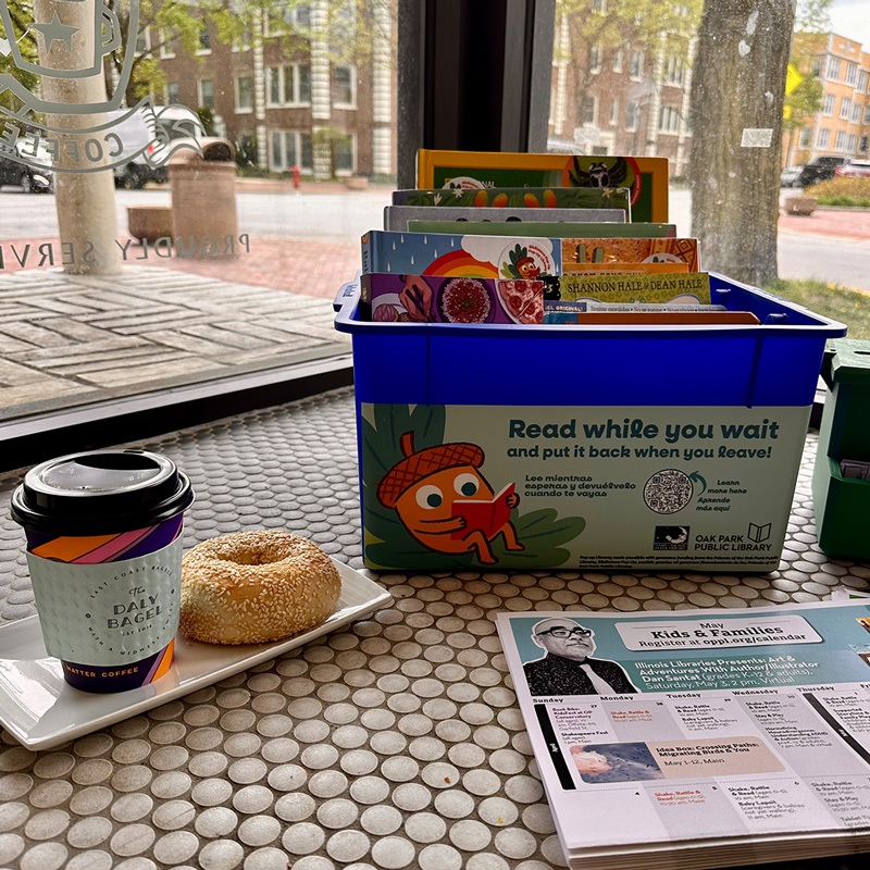 In a cafe window, there's a tray with a to-go coffee cup and a sesame seed bagel, a bin of kids books with a sign that says Read while you wait, and paper event calendars