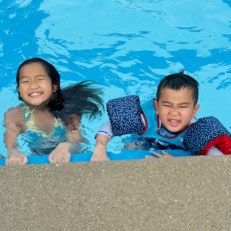 Two kids look up from the edge of a swimming pool, smiling and squinting into the sun