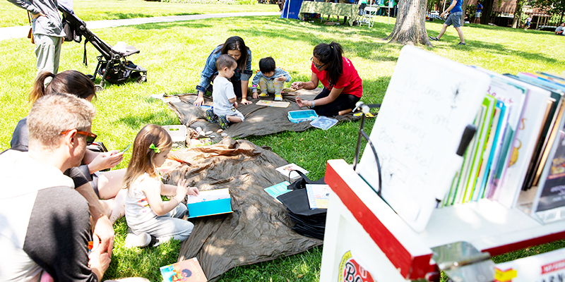 A library staff member helps two young children seated on a blanket in the grass select beads from a container; a book bike displaying children's books stands in front.