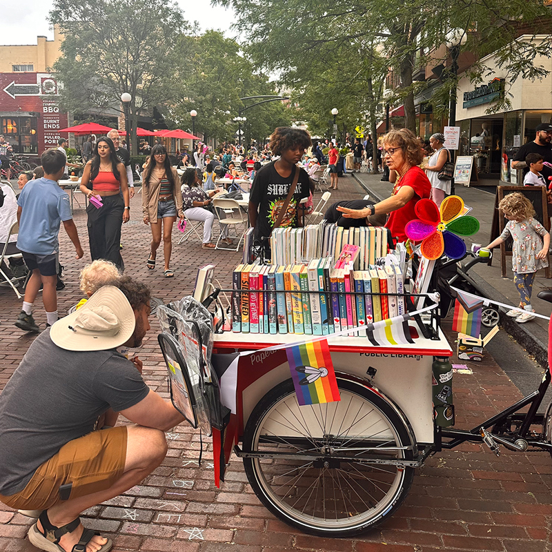 People engage with a cargo bike full of books on a street full of people and dining tables in summertime