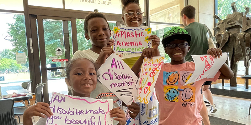 An adult and three kids all smile and hold up handmade signs in the Main Library entryway in support of disability pride and awareness.