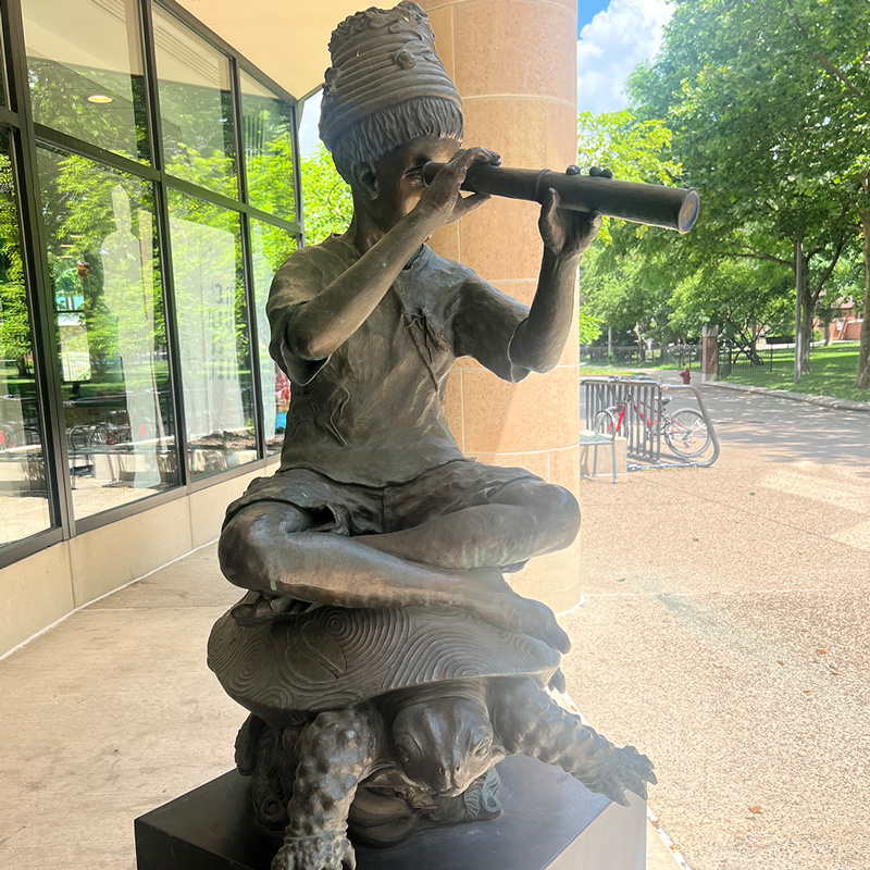 On the plaza outside the Main Library, a statue of a person sitting on top of a turtle and looking through a spyglass.