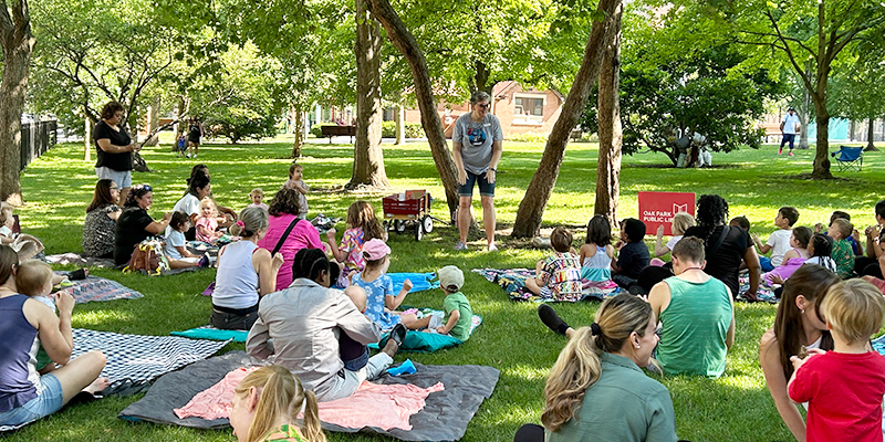 A library staff member stands in front of a group of families with young children sitting on picnic blankets under some trees in a park.