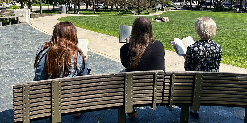 Three adults read books on a park bench on a sunny day.
