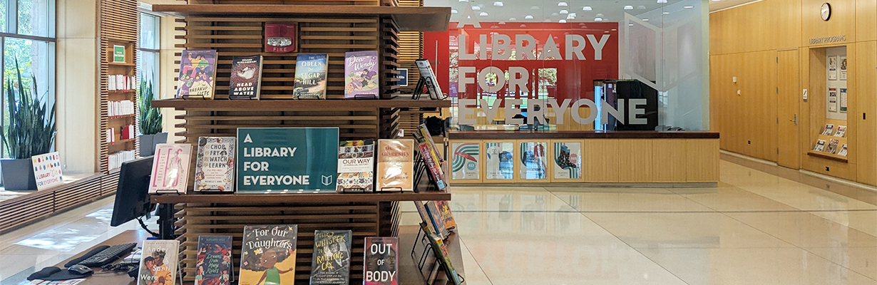 In the foreground, a books on display on a pillar next to a sign that reads A Library for Everyone; in the background, A Library for Everyone etched into a glass wall.
