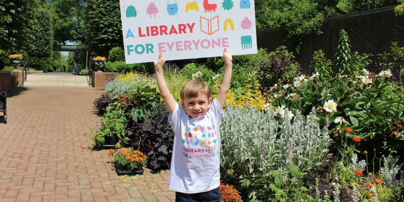 Child holds up oversized library card that reads A Library for Everyone, outside in a garden.