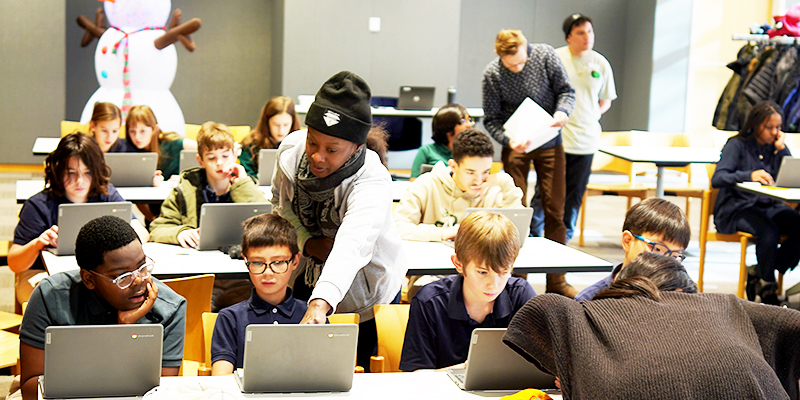 Students sit at tables with laptops in the library. An adult points to a young person's computer screen, and three other adults are walking around the room.