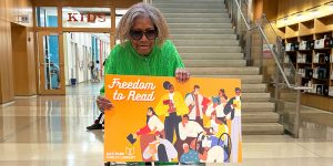 Person holding a large novelty library card propped up on their walker in front of the set of stairs inside the library