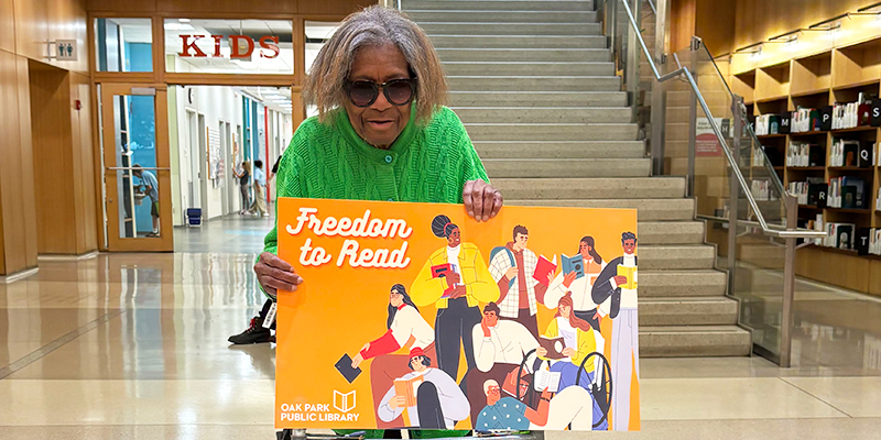 Person holding a large novelty library card propped up on their walker in front of the set of stairs inside the library