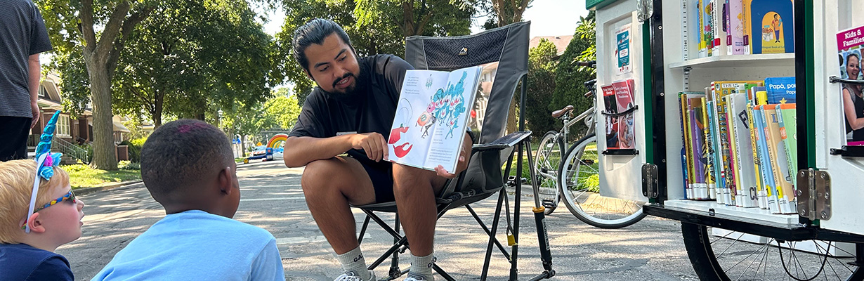 Library staff member reads a picture book to children at a block party. One of the library book bikes next to them is open to show lots of books and brochures.