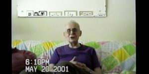 A screenshot from a home video. An older man with glasses sits on a couch facing the viewer, smiling and appearing to be midsentence. The date May 20, 2001 appears on the lower left.