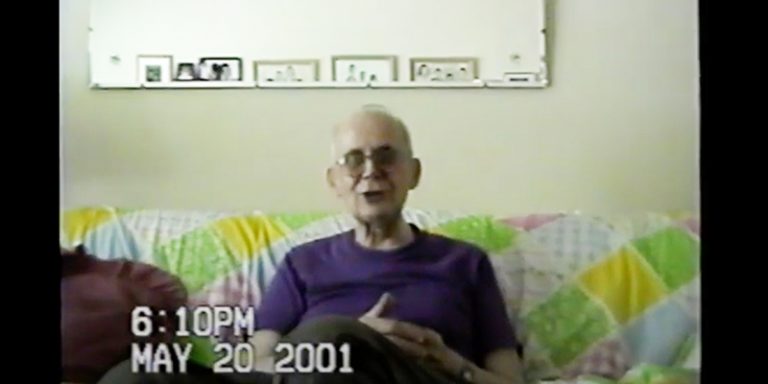 A screenshot from a home video. An older man with glasses sits on a couch facing the viewer, smiling and appearing to be midsentence. The date May 20, 2001 appears on the lower left.