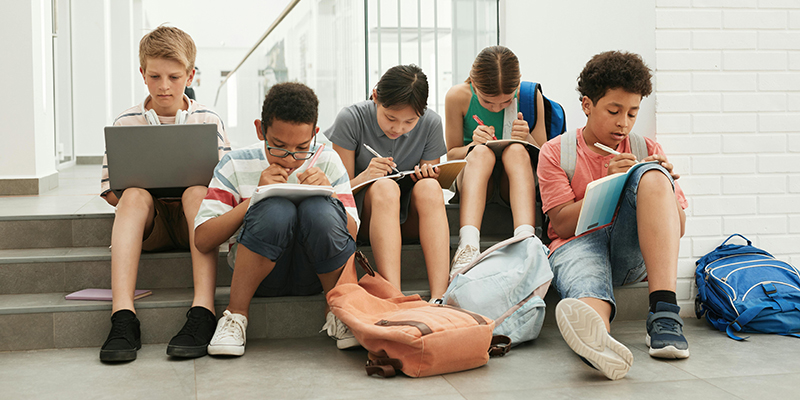Students sitting on steps in a school hallway, using a laptop, and writing in notebooks.