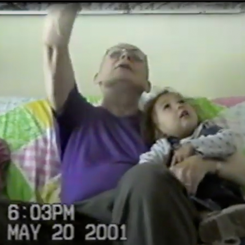 A screenshot from a home video. An older man holds a young girl on the couch, appearing to be telling her a story as he points upward. The date May 20, 2001 appears on the lower left.