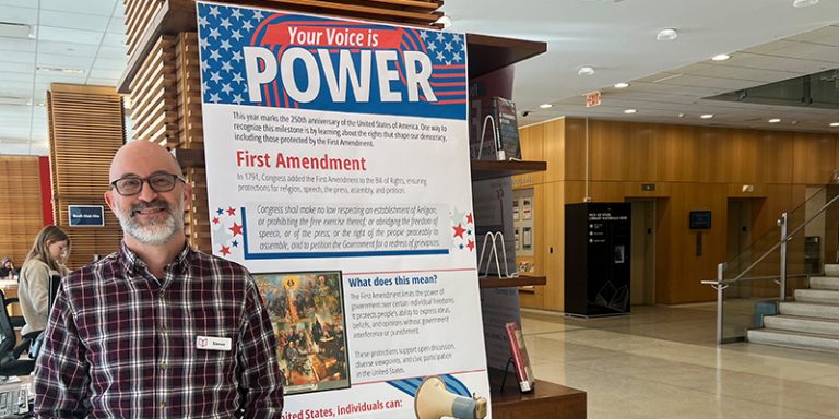 Simon Czerwinskyj stands in front of a library display. A poster reads Your Voice Is Power and has information on the First Amendment.