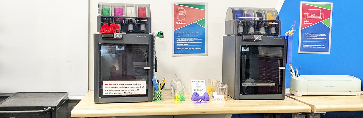 Two 3D printers sent on a tabletop with 3D-printed items displayed in between.