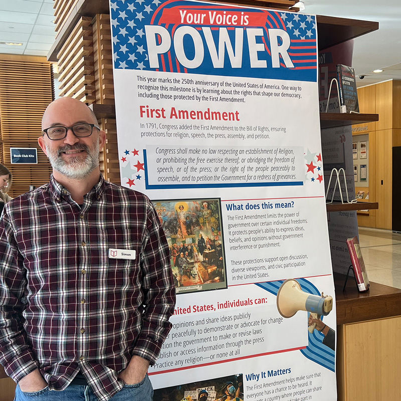 Simon Czerwinskyj stands in front of a library display. A poster reads Your Voice Is Power and has information on the First Amendment.