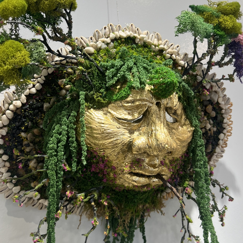 Mixed media 3D artwork of face surrounded by trees, moss, and pebbles.