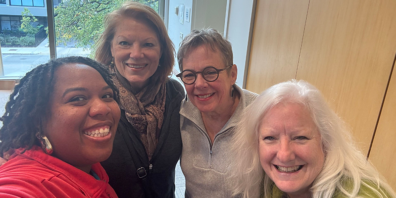 Four women group together and smile for a selfie.