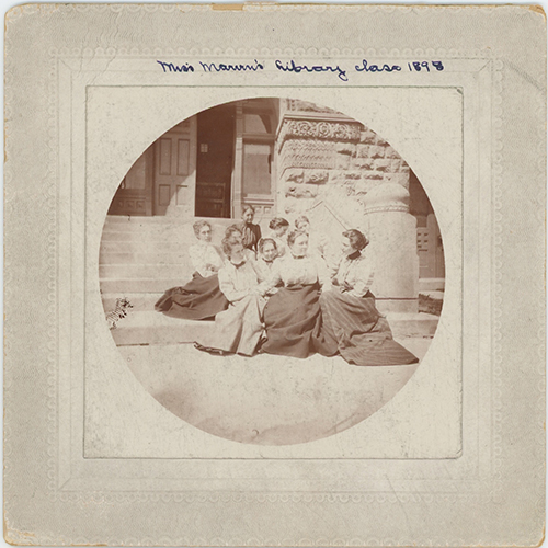 Photo of women sitting on stairs in front of a building with the words Miss Maureen's library class 1898 written above.