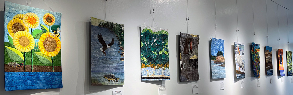 Art quilts hang on the wall of a gallery.