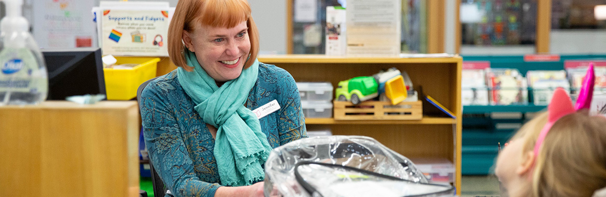 Librarian hands a backpack to a child at a library service desk.