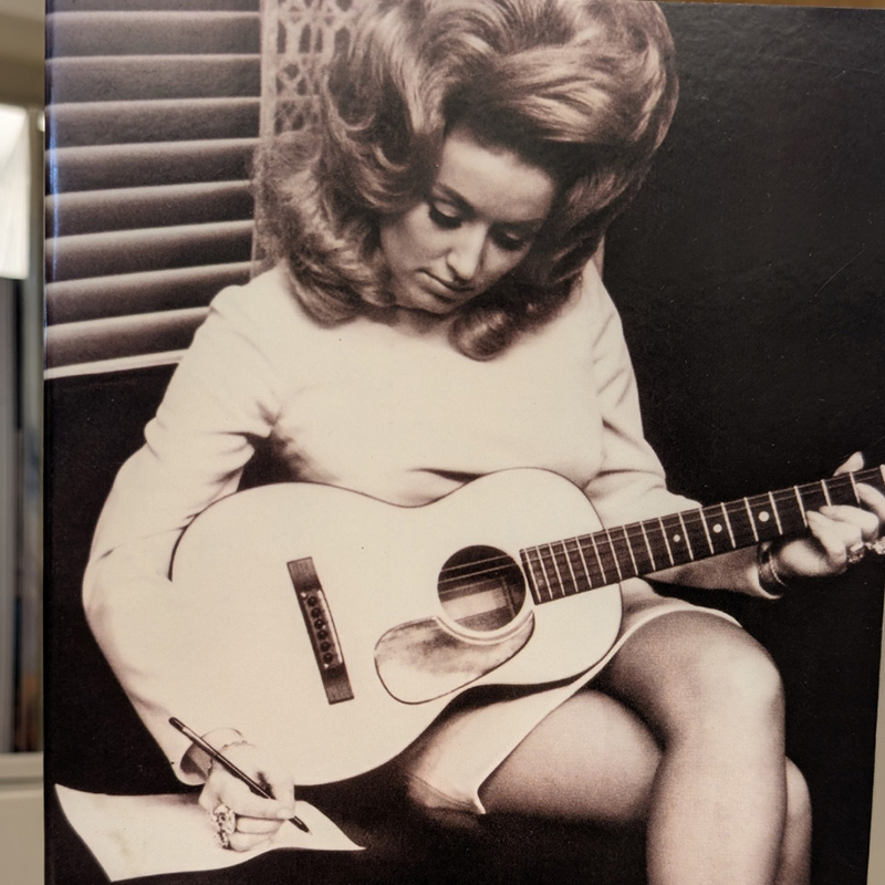 Dolly Parton with guitar, writing on a piece of paper. From book cover of Unlikely Angel: The Songs of Dolly Parton.