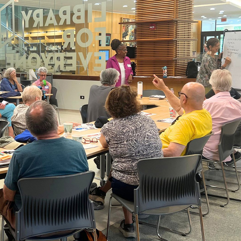 Hands are raised among people at tables in the library, as one person stands at the front and another writes on a whiteboard.